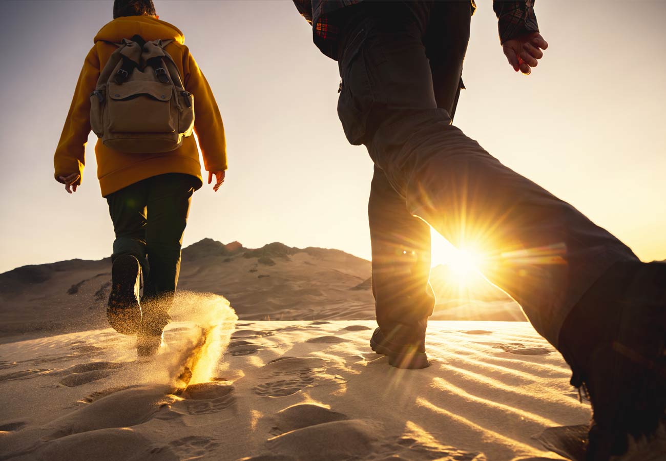 Two hikers with backpacks walks in sunset desert dunes.