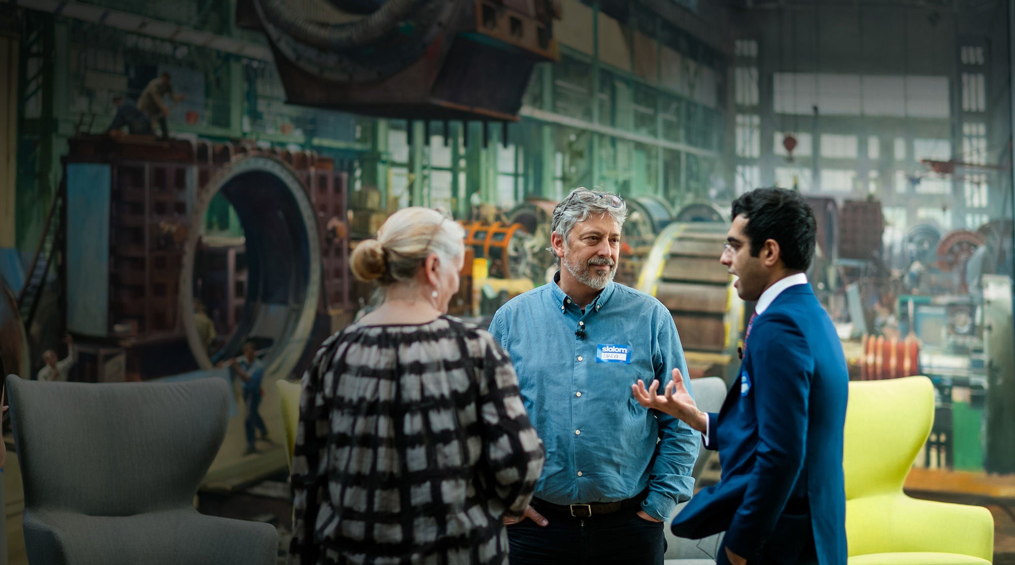 Man in blue button-down with Slalom name tag reading "Luca" speaking to a man in a blue suit jacket and a woman in a black and gray printed top.