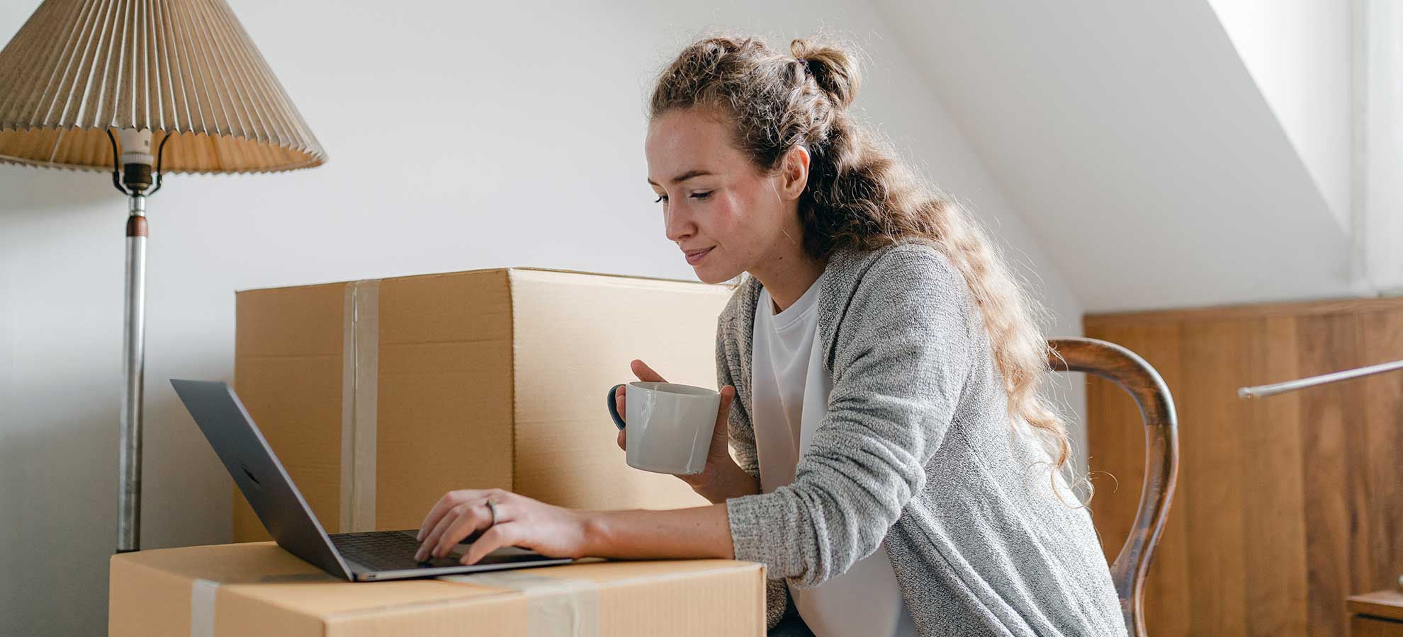 A woman sits among moving boxes typing on a laptop.