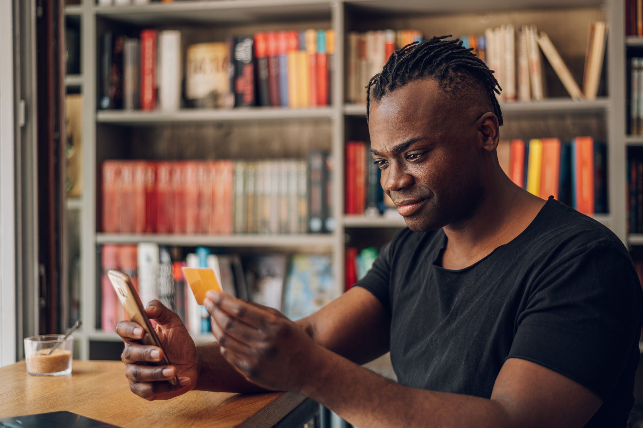 Portrait of an african american male holding credit card and using smartphone in a cafe. Using online banking application on his mobile phone. Young freelancer receive payment, check balance.