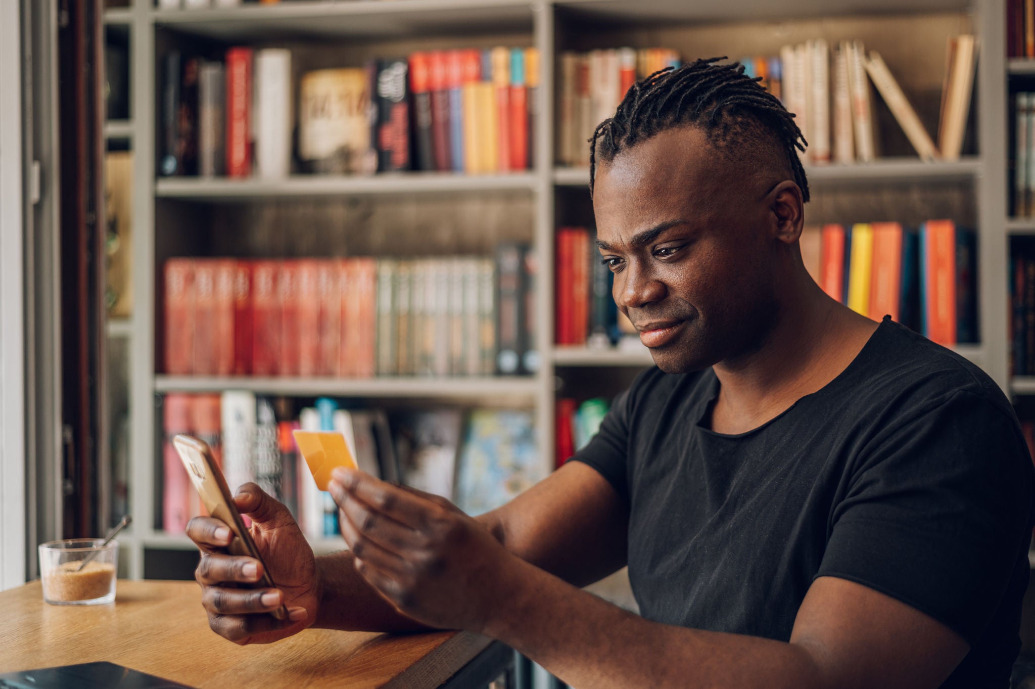 Portrait of an african american male holding credit card and using smartphone in a cafe. Using online banking application on his mobile phone. Young freelancer receive payment, check balance.