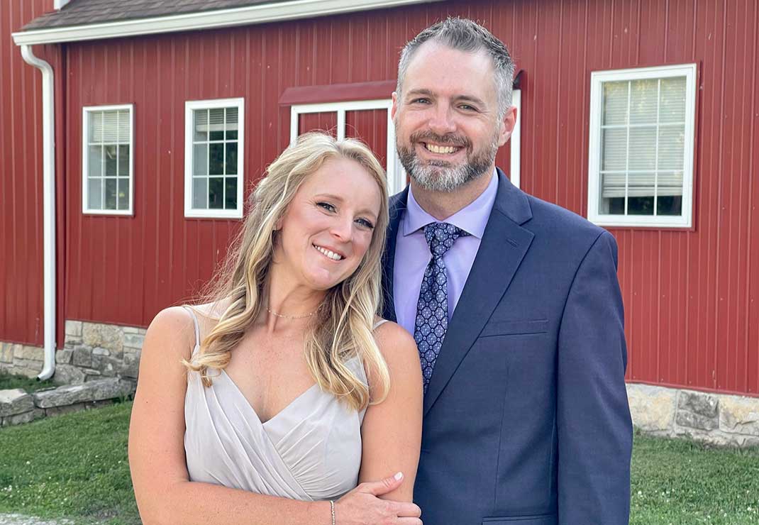 General manager Saint Fults wearing a gray suit jacket, lavender shirt, and printed tie, standing next to his wife in a silver dress in front of a red barn.