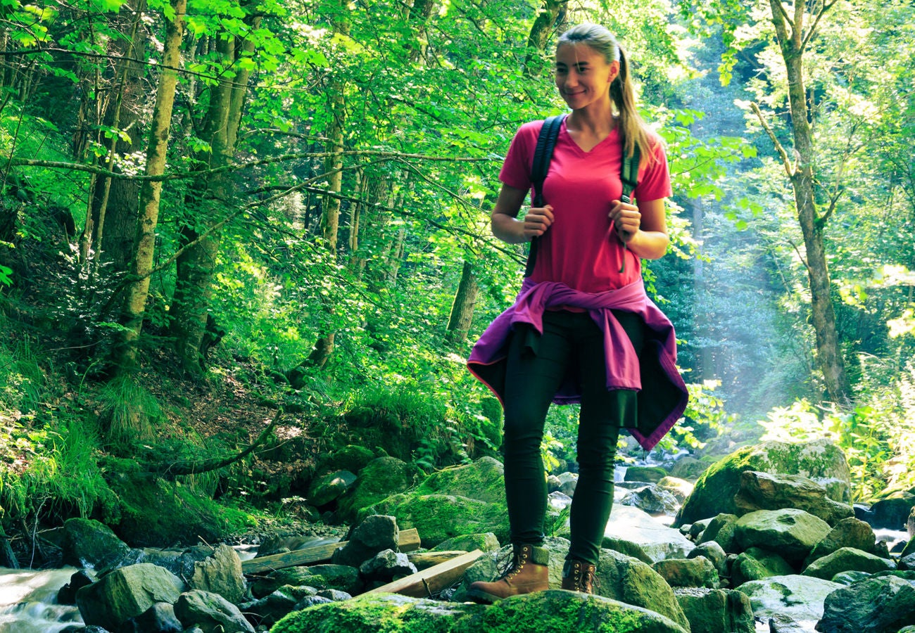 A woman walks near a stream in a green forest during the day.