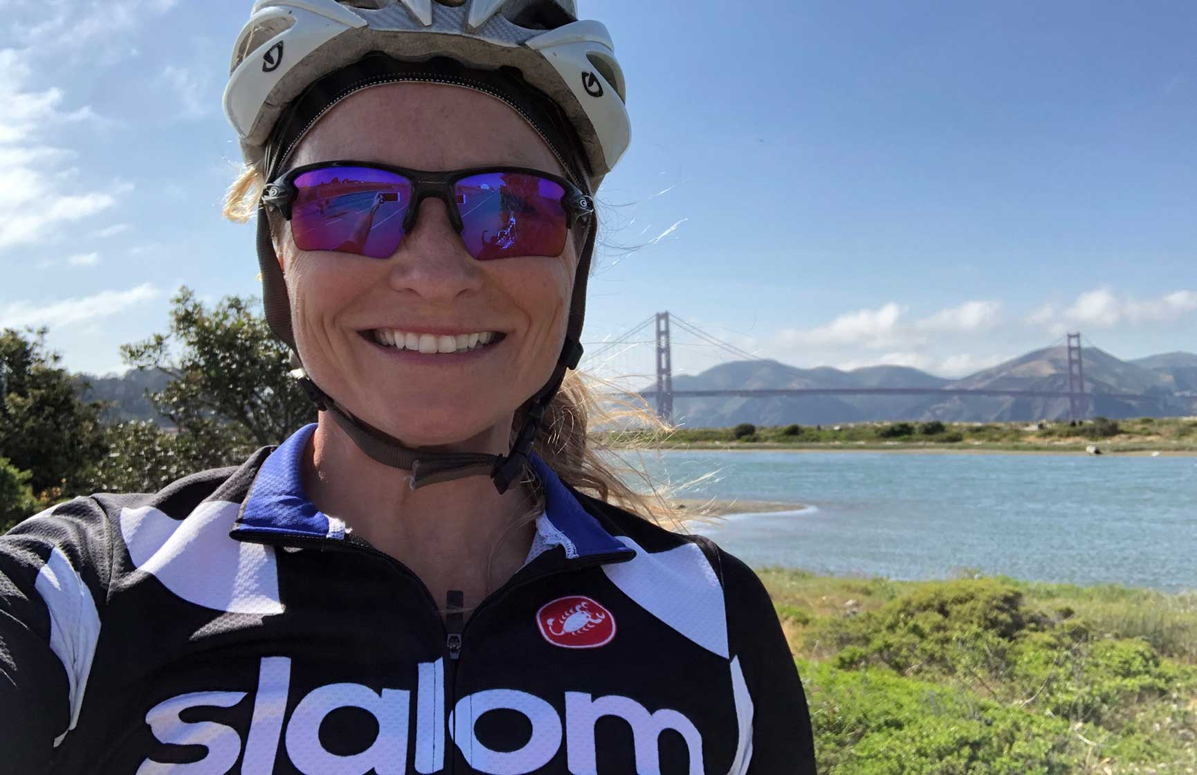 NorCal general manager Sarah Duffy wearing a helmet, sunglasses, and black and white slalom bike shirt, posing in front of the San Francisco Bay with the Golden Gate Bridge in the distance.