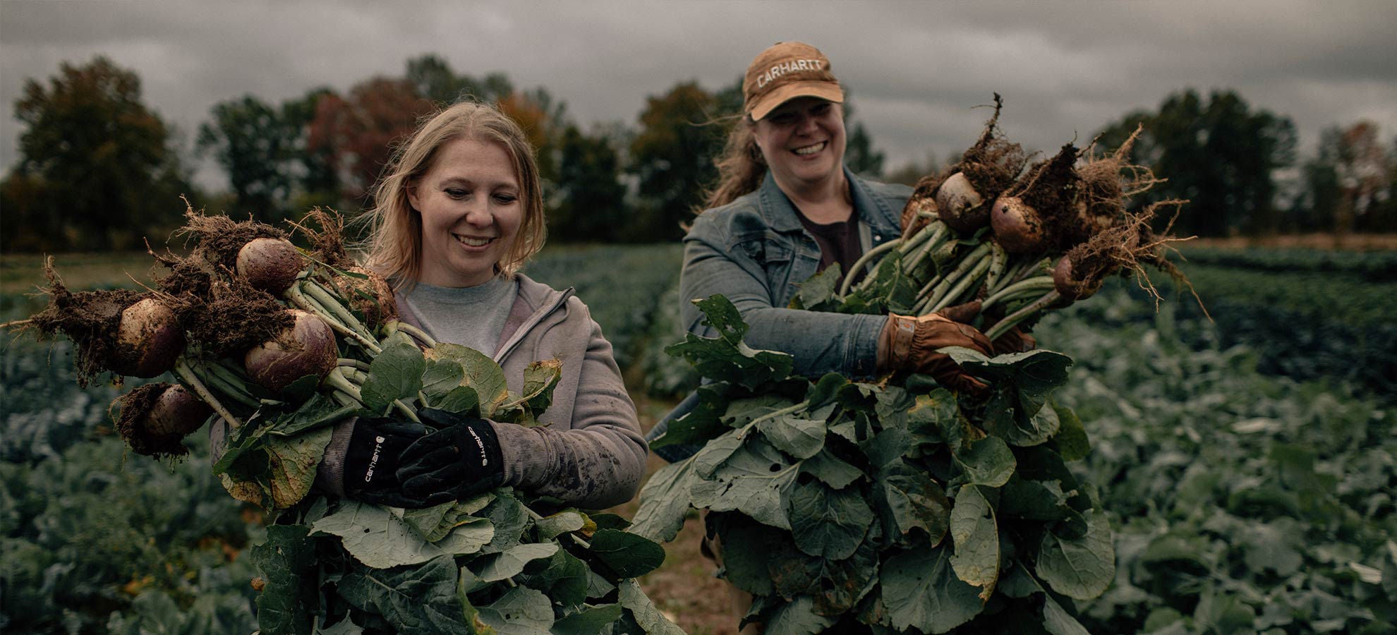 Two female farmers holding bundles of fresh produce in field.