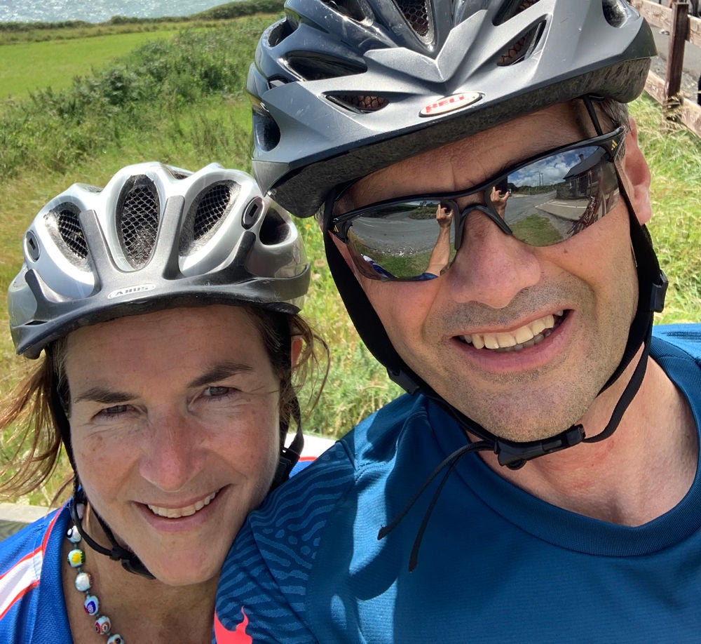 Ireland general manager Jane Dawson, wearing a silver biking helmet, standing next to her husband in a silver biking helmet, sunglasses, and blue shirt.