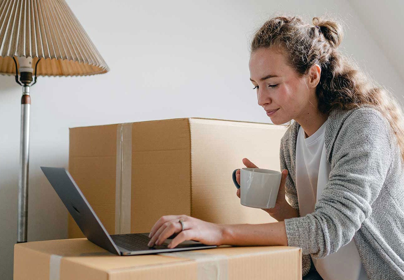 Woman uses a laptop and holds a cup of coffee surrounded by moving boxes.