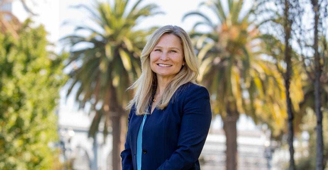Headshot of NorCal's general manager, Sarah Duffy, smiling and wearing a blue shirt and blazer against a backdrop with two palm trees.