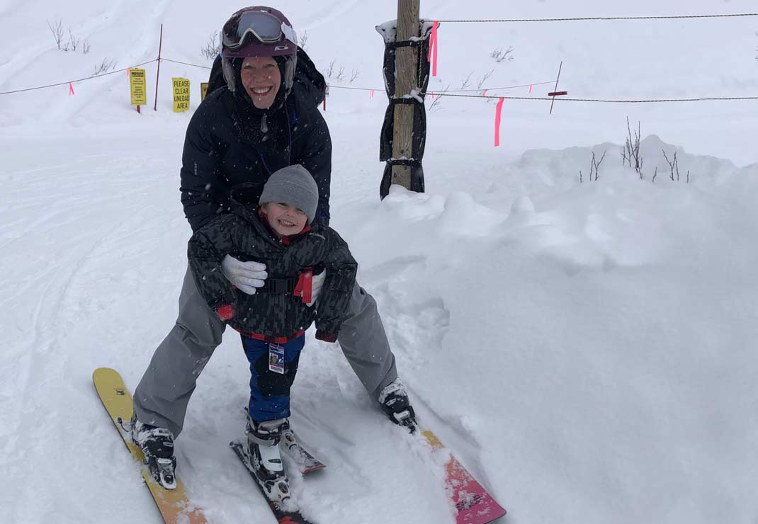 Salt Lake City general manager, Isla Bragg, in black and gray ski gear and a purple helmet, smiling and holding her son as he skis between her legs.