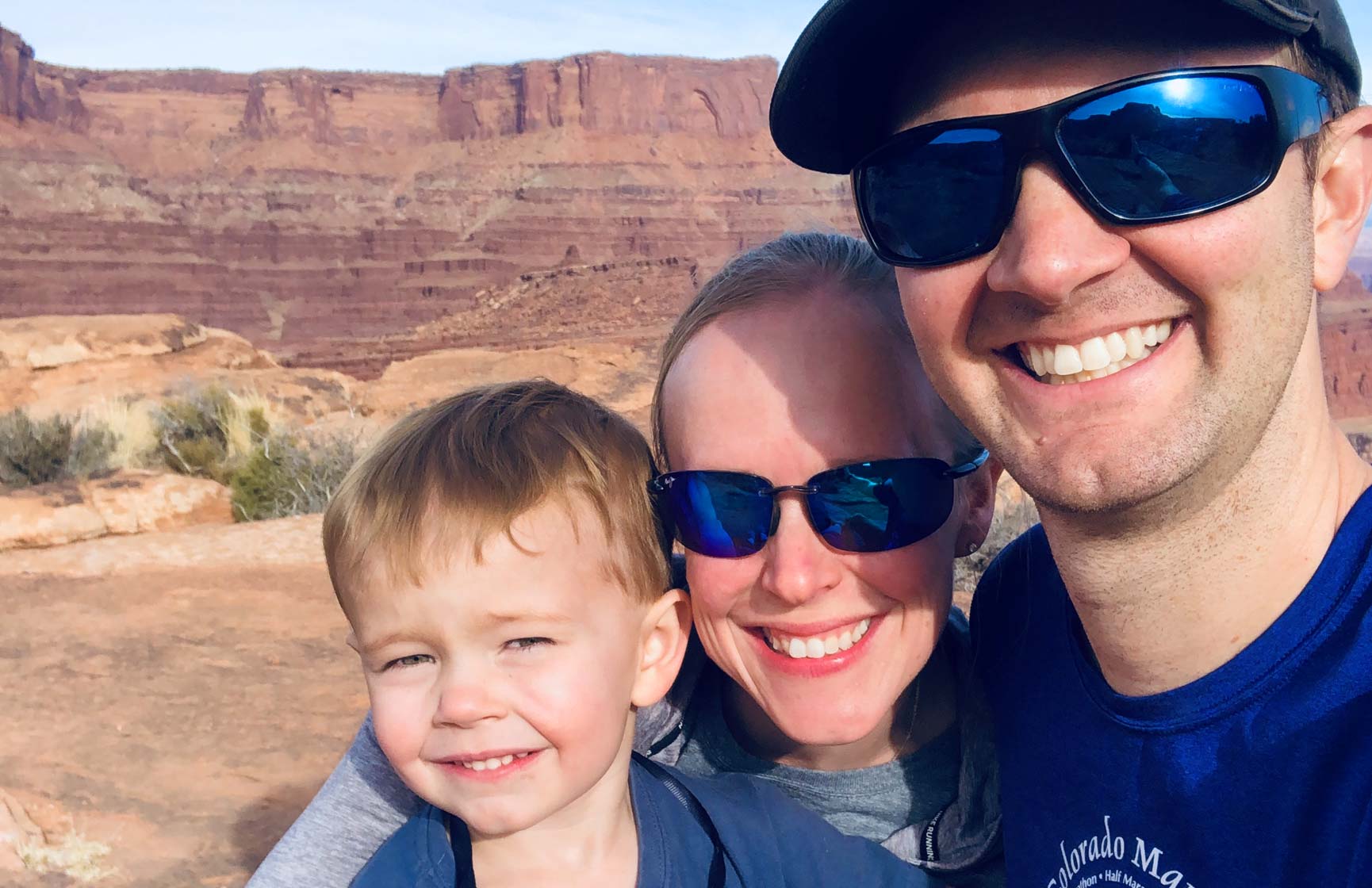 Salt Lake City general manager Isla Bragg and her husband, both wearing sunglasses, posing with their son against a red, mountainous backdrop.