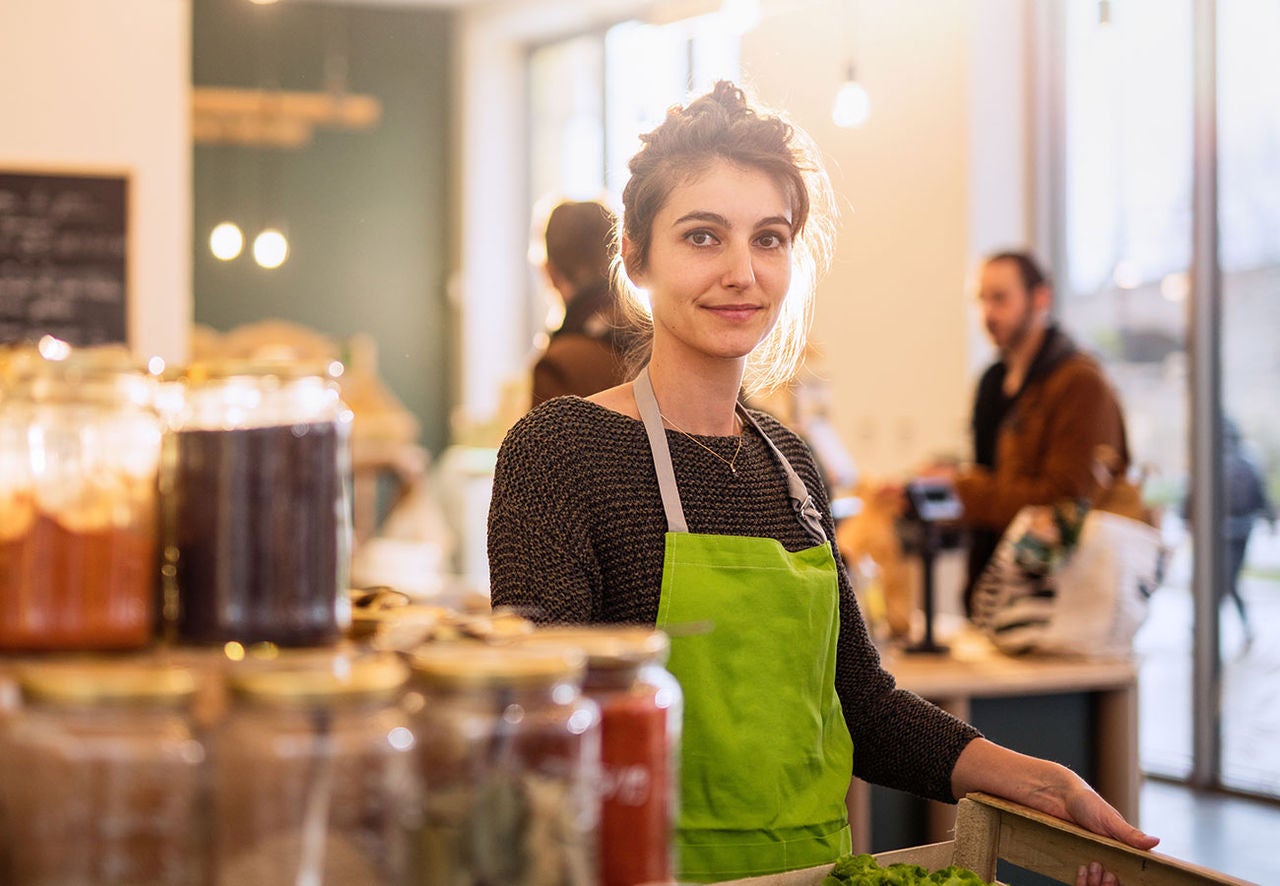 Young woman working in a bulk food store with a crate of salads in her hands.