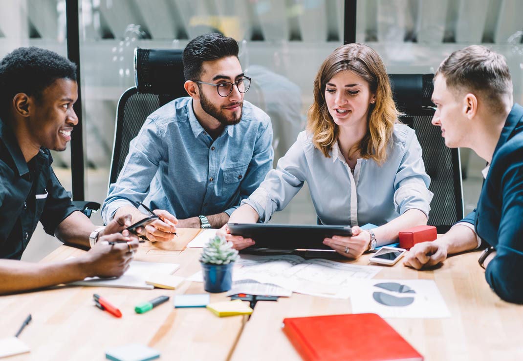 A diverse group of four employees sitting around a table collaborating on a digital tablet.