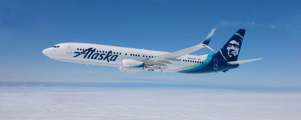 Alaska Airlines airplane flying above the clouds in a clear blue sky.