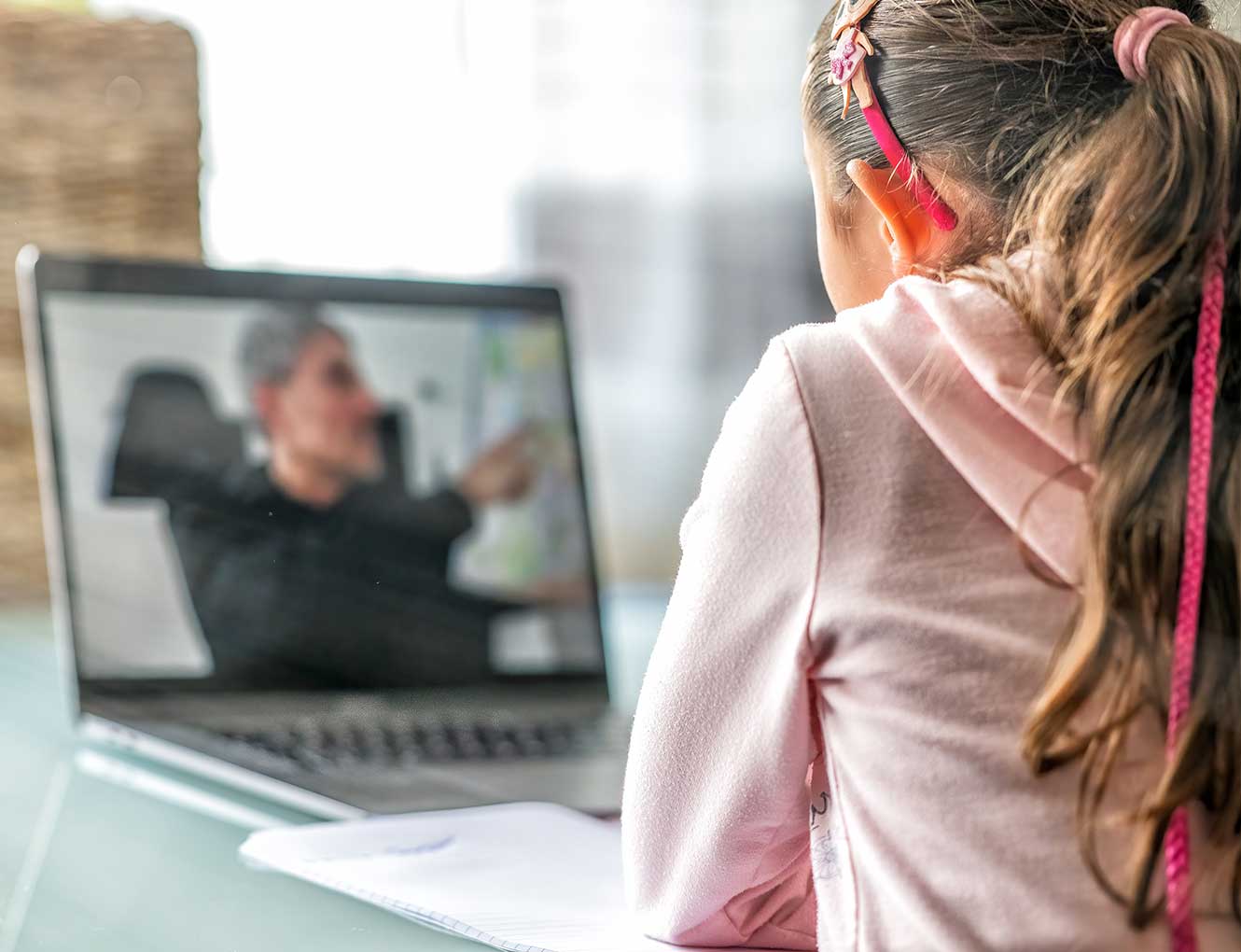 A young girl taking notes as she watches her teacher on her laptop.