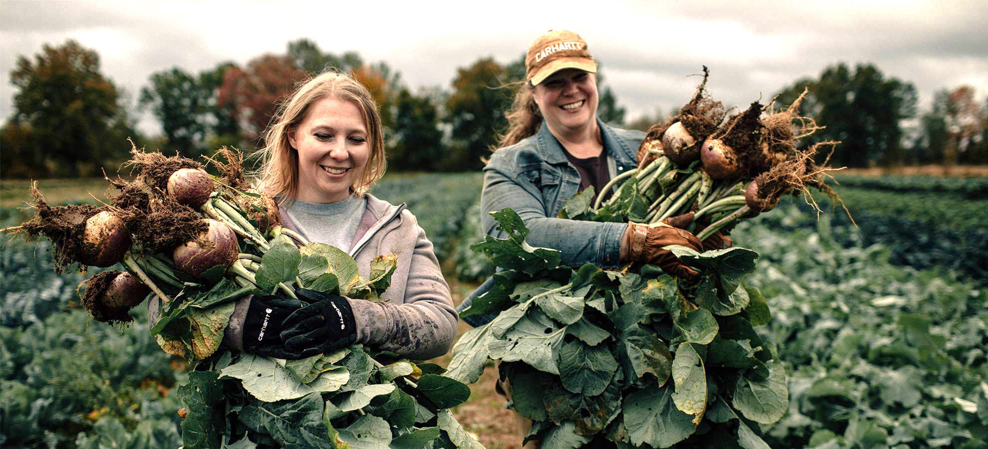 Two female farmers holding bundles of fresh produce in field.