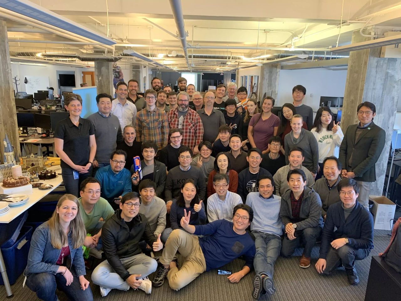 A large group of coworkers pose together for a team photo in an open-plan office. People of various ages and backgrounds are smiling and sitting or standing in several rows. The workspace features desks, computers, exposed ceilings, and concrete pillars. A table with cakes and refreshments is visible on the side, suggesting a celebration or special event.